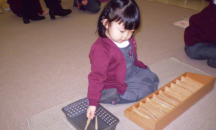 Create a realistic image of a diverse classroom with Montessori materials, showing a young Asian girl confidently working independently with a wooden puzzle, while other children of various ethnicities engage in their own activities in the background, emphasizing a bright, organized, and nurturing environment that promotes self-reliance and independence.